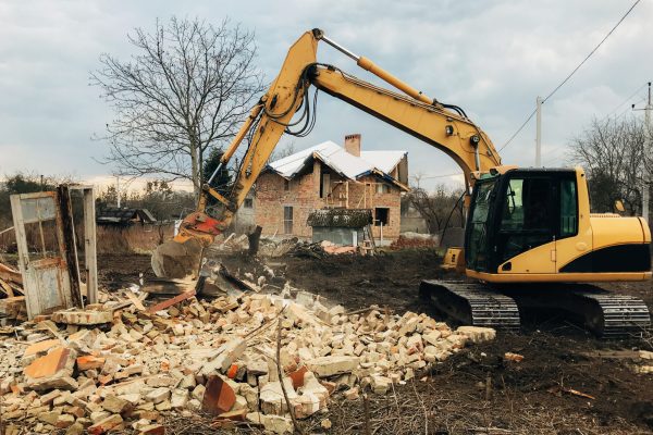 Excavator destroying brick house on land in countryside. Bulldozer clearing land from old bricks and concrete from walls with dirt and trash. Backhoe machinery ruining house